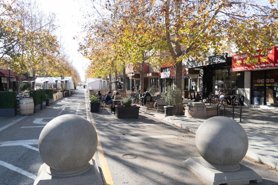 Photo of a downtown area where cars are no longer allowed on the street so restaurant seating is placed in parking spaces and vehicle lanes. It is a sunny day with people sitting outside